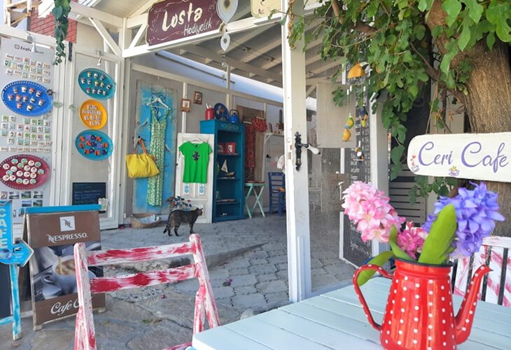 Selimiye&rsquo;de Losta Hediyelik ve Ceri Cafe&rsquo;nin &ouml;n&uuml;nde, mavi masada &ccedil;i&ccedil;ekli s&uuml;rahi, renkli sandalyeler ve fonda el işi &uuml;r&uuml;nlerin sergilendiği d&uuml;kk&acirc;n girişi, arka planda bir kedi.