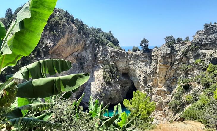 Gazipaşa Delikdeniz Kral Koyu, &ouml;n planda muz yaprakları ve yeşillikler arasında, kayalıklar ve doğal mağarayla birlikte g&ouml;r&uuml;l&uuml;yor.